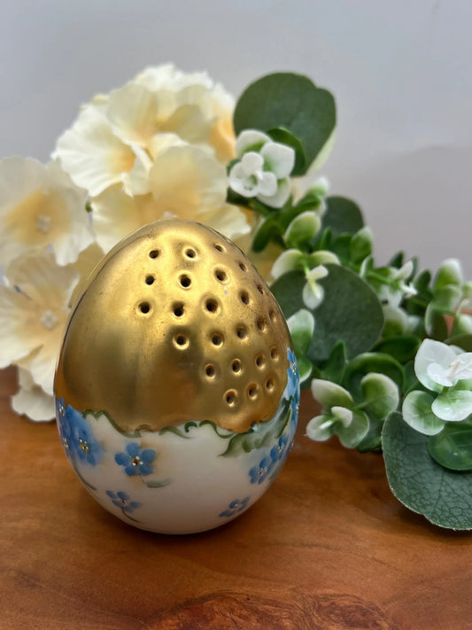 Decorative china egg shaker with floral design and gold top on a wooden surface with flowers in the background