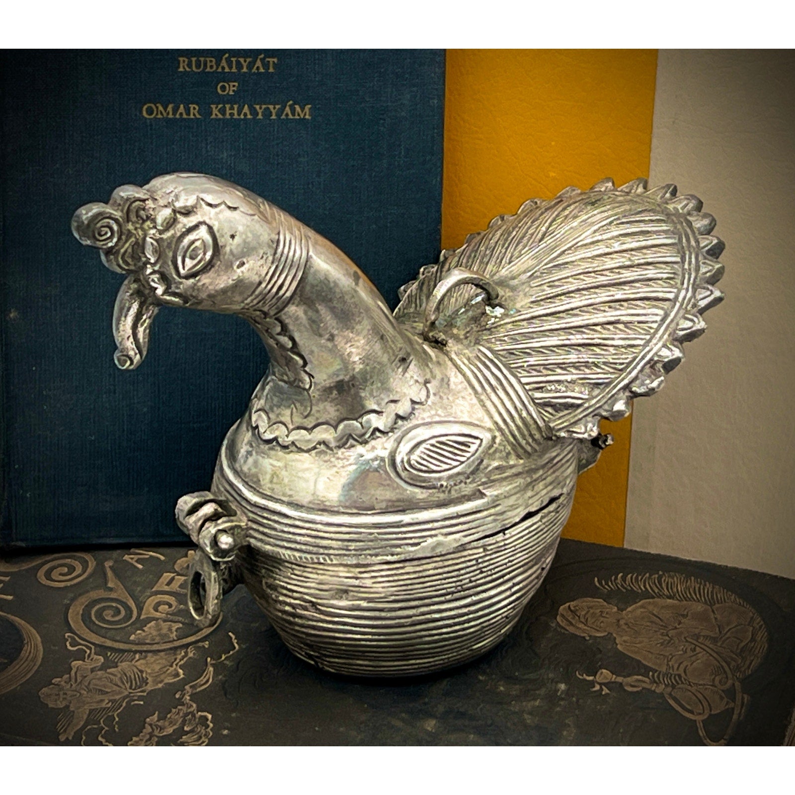 Decorative silver bird-shaped container on a textured surface with a book titled 'Rubaiyat of Omar Khayyam' in the background.