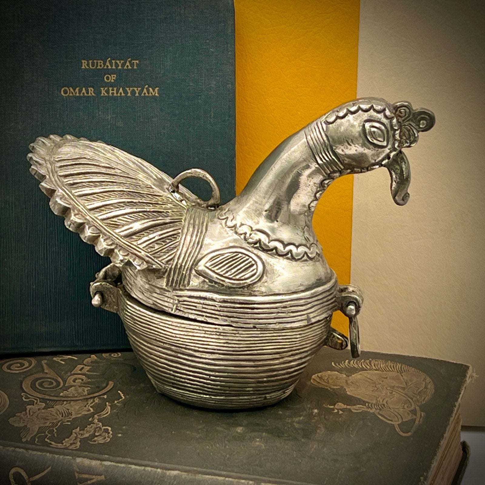 Decorative silver bird-shaped object on a stack of books with a blurred book titled 'Rubáiyát of Omar Khayyám' in the background.